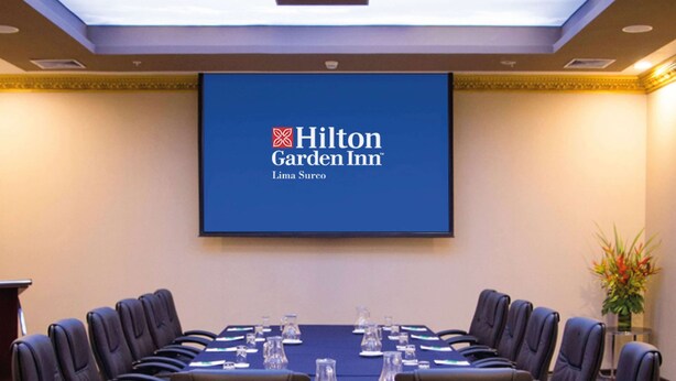 Une salle de conférence avec une longue table, des chaises noires et un écran affichant le logo de l’hôtel Hilton Garden Inn.