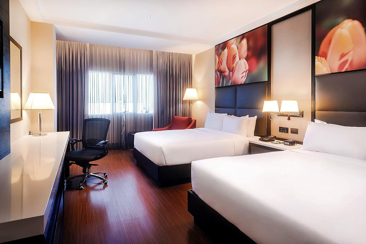A hotel room with two beds, a desk, a chair, a lamp, and a large window with curtains.