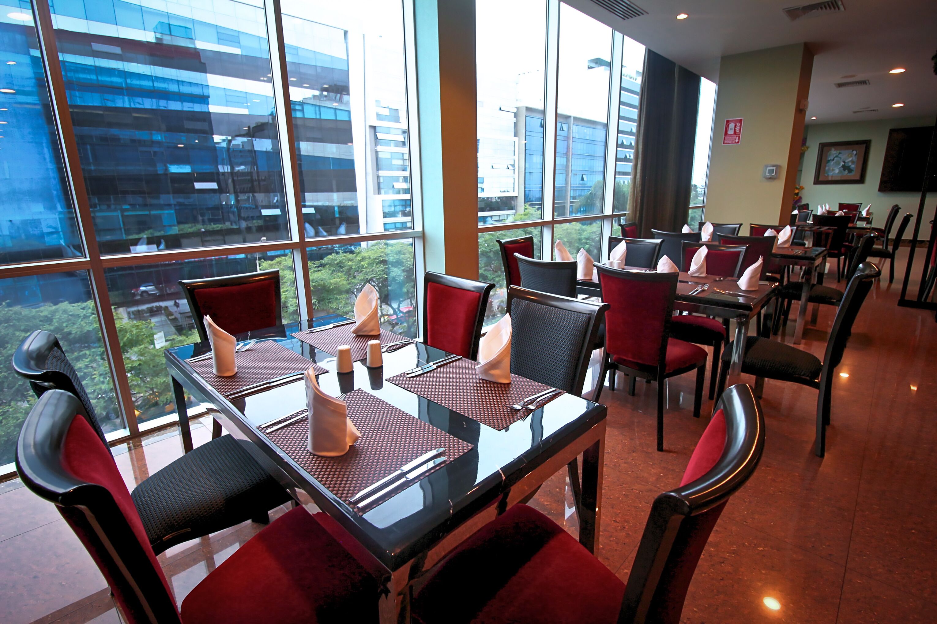 Un restaurante con mesas dispuestas para comer, ventanas amplias que ofrecen vistas a edificios y sillas con cojines rojos.