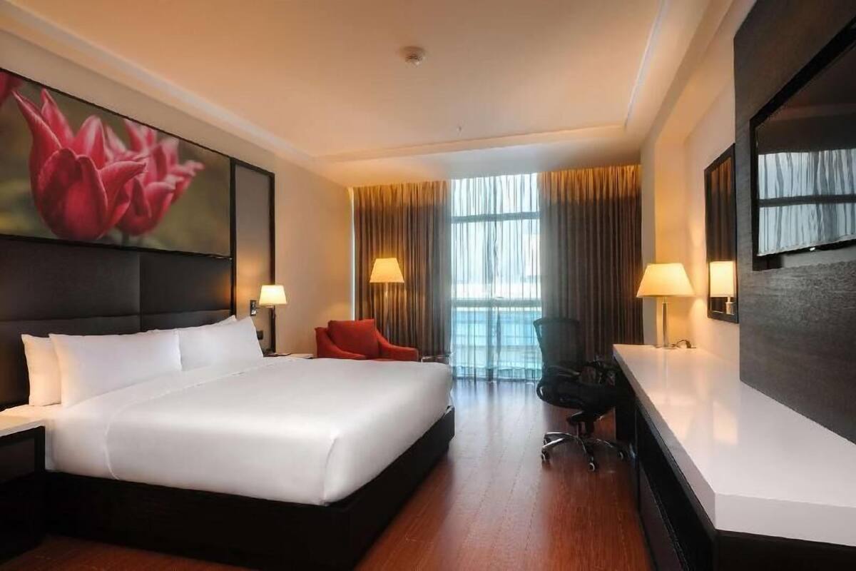 A hotel room with a large bed, two bedside tables, a desk with a chair, a large window with curtains, and a wall-mounted TV.