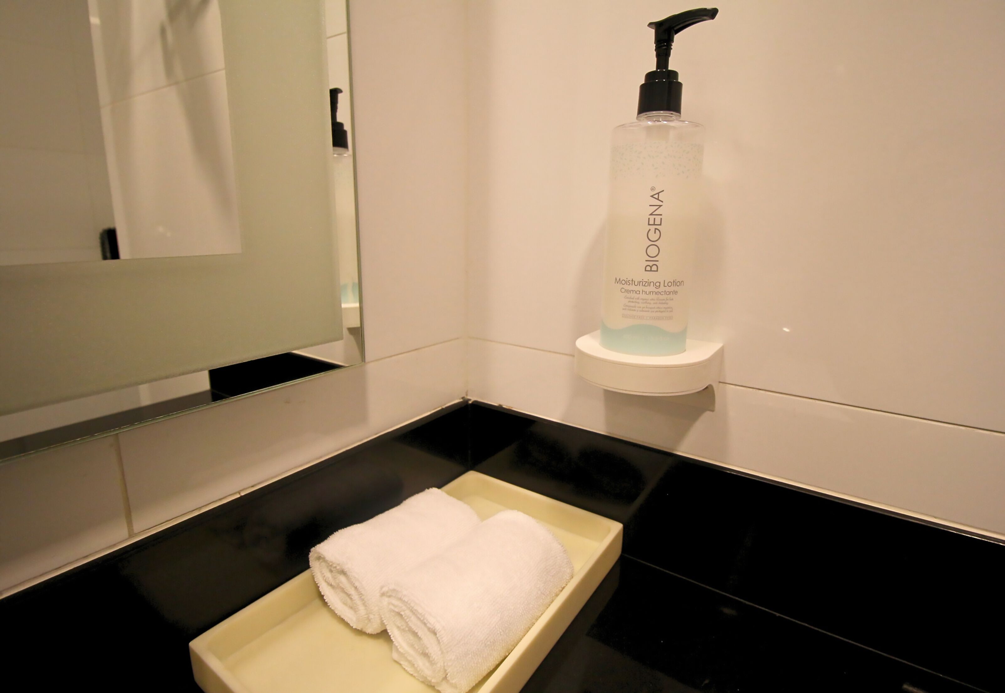 Uma casa de banho com bancada preta, uma toalha branca sobre um porta-toalhas e um frasco de Loção Hidratante Biogena numa estante branca.