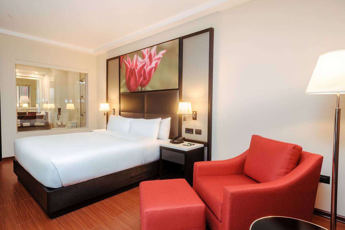 A hotel room with a large bed, a red armchair, a nightstand, and a wall-mounted TV.