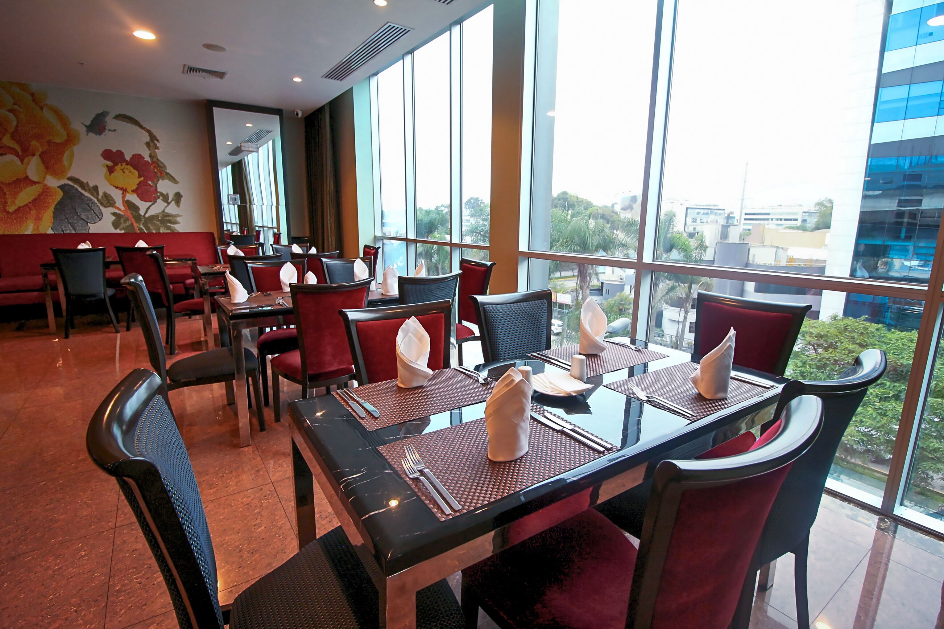 Un restaurante con mesas dispuestas para comer, amplios ventanales que ofrecen vistas a la ciudad y un mural en la pared.