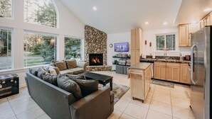 A modern living room with a stone fireplace, a large window, and a kitchen with wooden cabinets and a stainless steel refrigerator. - 01sl - Hot Tub - Pet Friendly - Bbq - Sleeps 8 3 Bedroom Home by RedAwning (Deming)