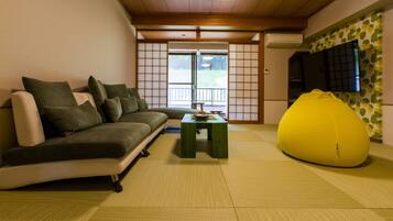 A modern living room with a sofa, a small table, a bean bag, tatami mats, a TV, and a sliding door leading to an outdoor area.
