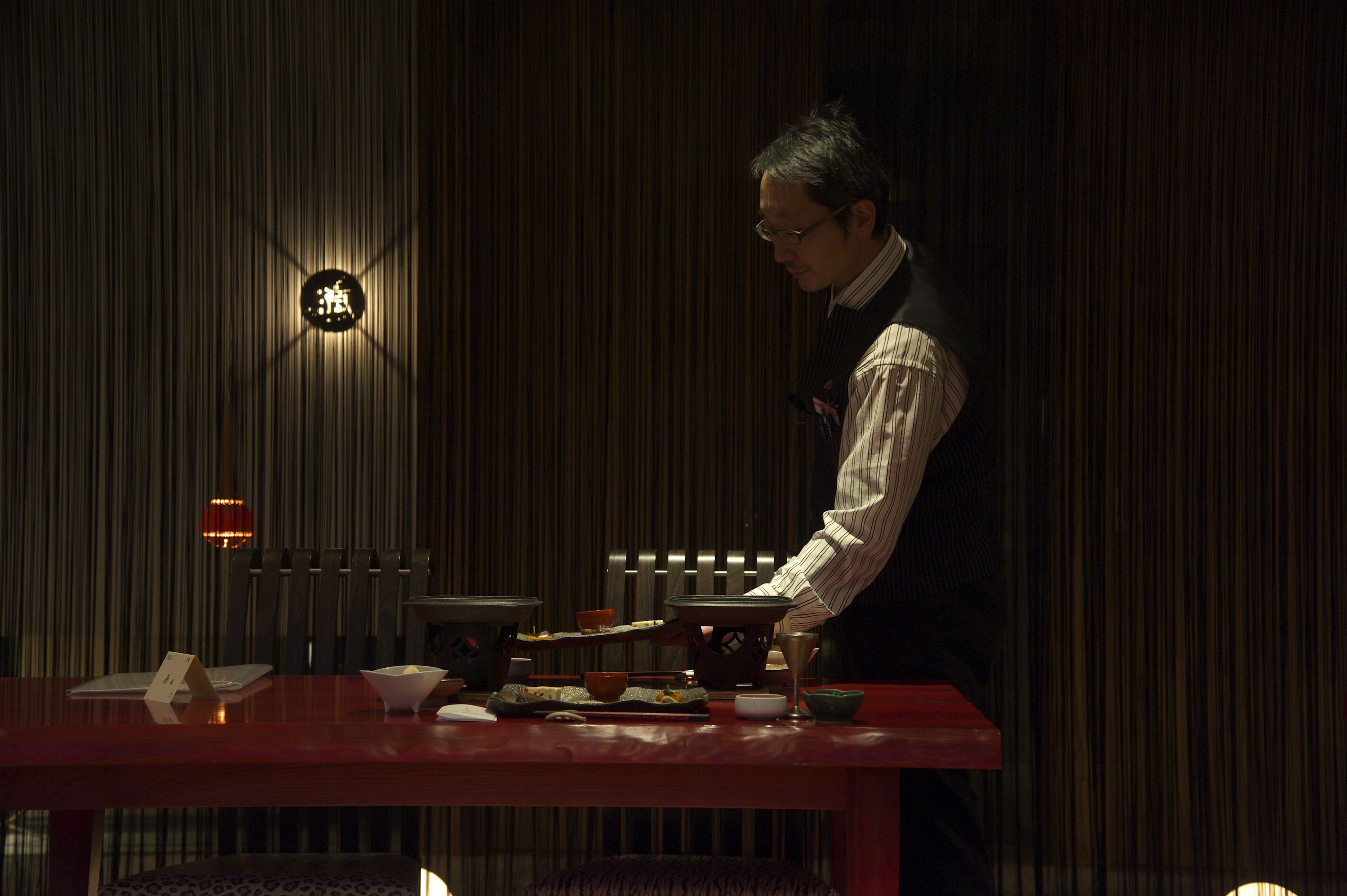A person serving food at a table with traditional dishes, a sign with Chinese characters, and a dimly lit room with vertical lines on the wall.