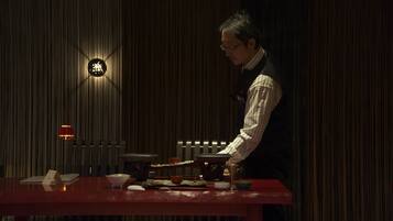 A person serving food at a table with traditional dishes, a sign with Chinese characters, and a dimly lit room with vertical lines on the wall.