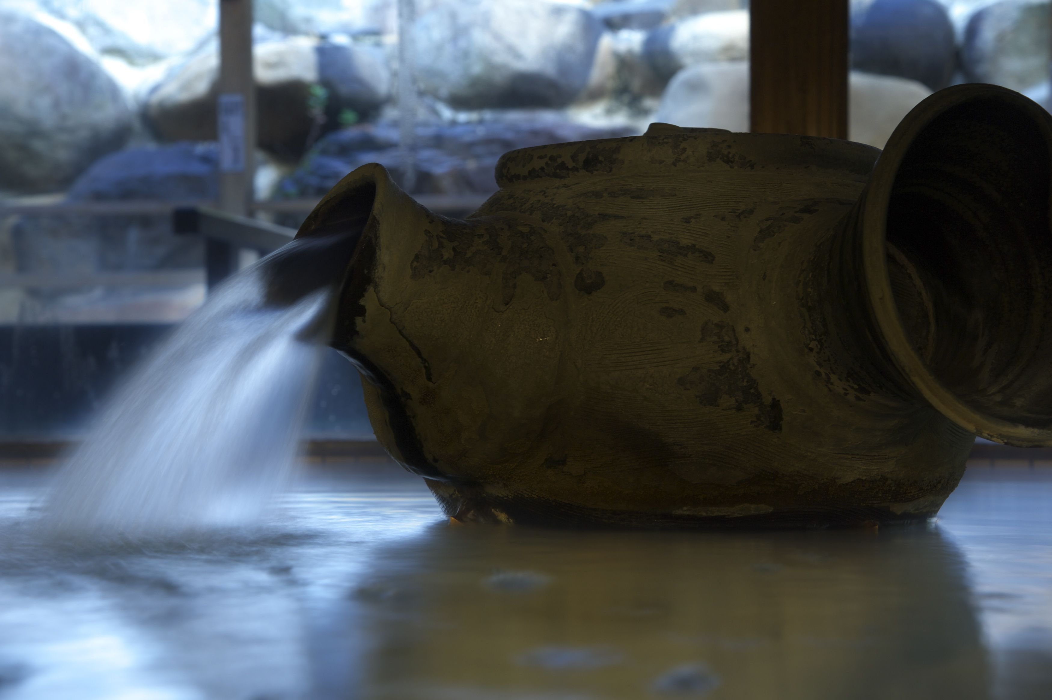 A rustic, weathered ceramic pot with a spout, positioned indoors with a blurred background of rocks and a wooden structure.