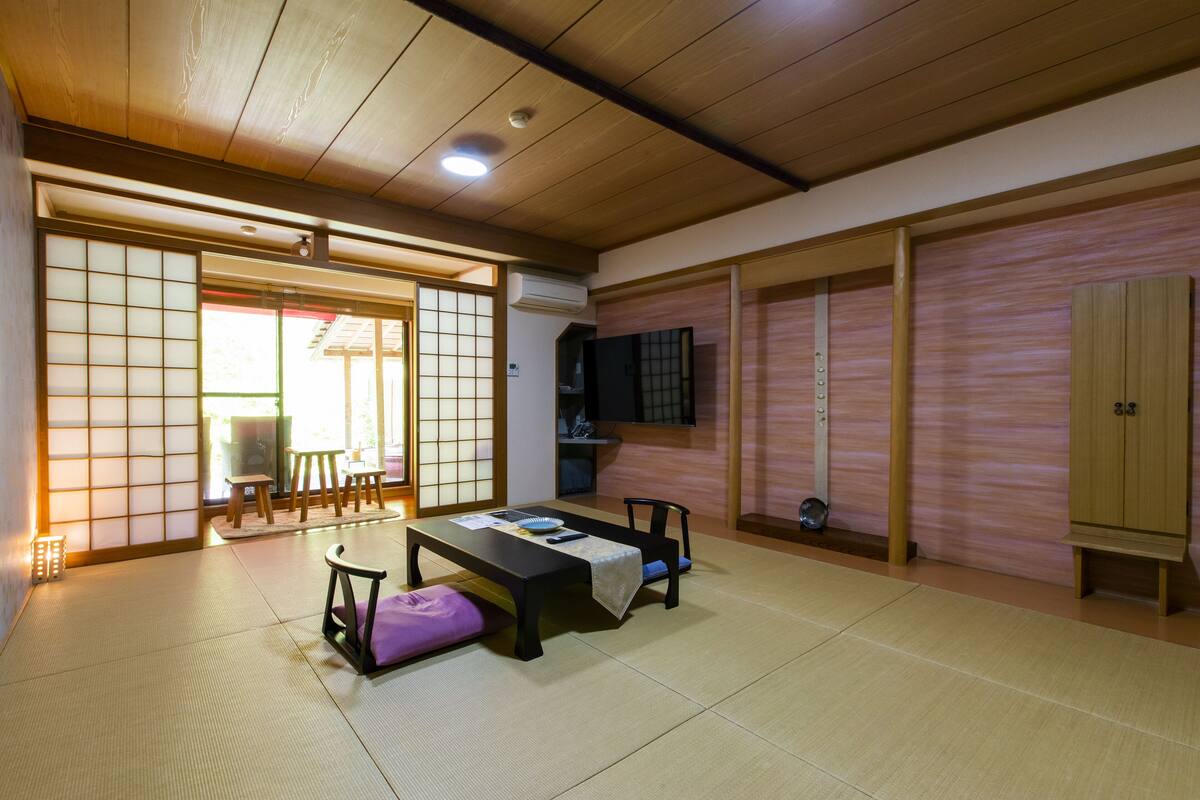 Ett traditionellt japanskt rum med tatamimattor på golvet, ett lågt bord med kuddar, en skjutdörr och en tv som är monterad på väggen.