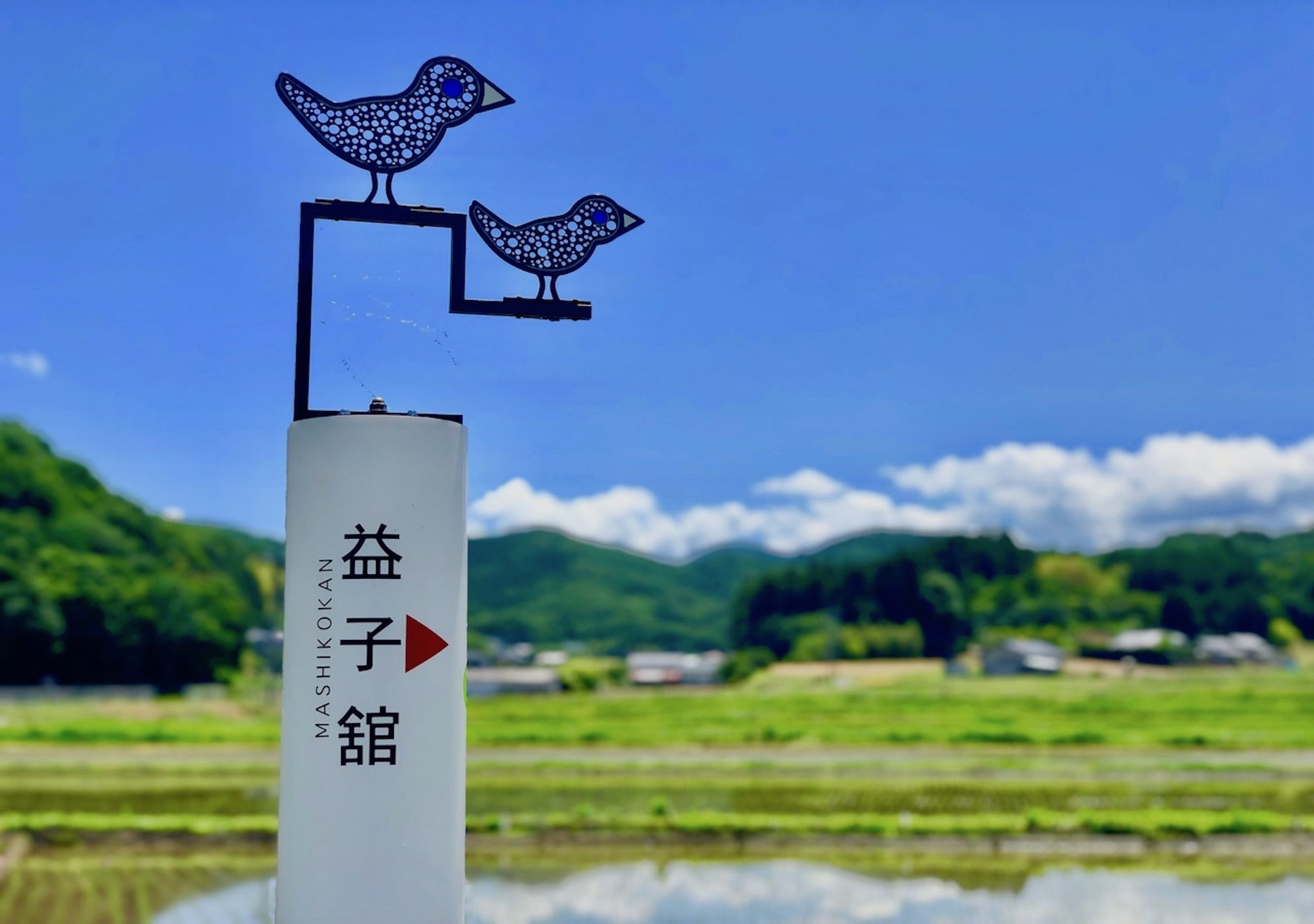 A directional sign with two stylized bird figures and Japanese characters, set against a rural landscape with fields and mountains.