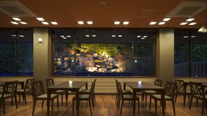 A dining area with wooden tables and chairs, large windows with a view of a rock garden, and recessed ceiling lighting.