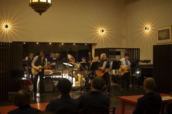 Una banda actuando en un escenario con instrumentos musicales y partituras, un público sentado al frente y un amplio ventanal al fondo.