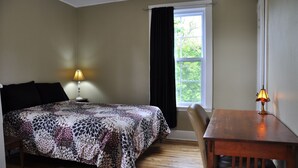 A bedroom with a bed, a bedside table with a lamp, a window with curtains, and a wooden chair. - Moncton Suites - 81 Maple Street (Moncton)