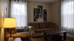 A living room with a sofa, a table, a lamp, and decorative wall art. - Moncton Suites - 81 Maple Street (Moncton)