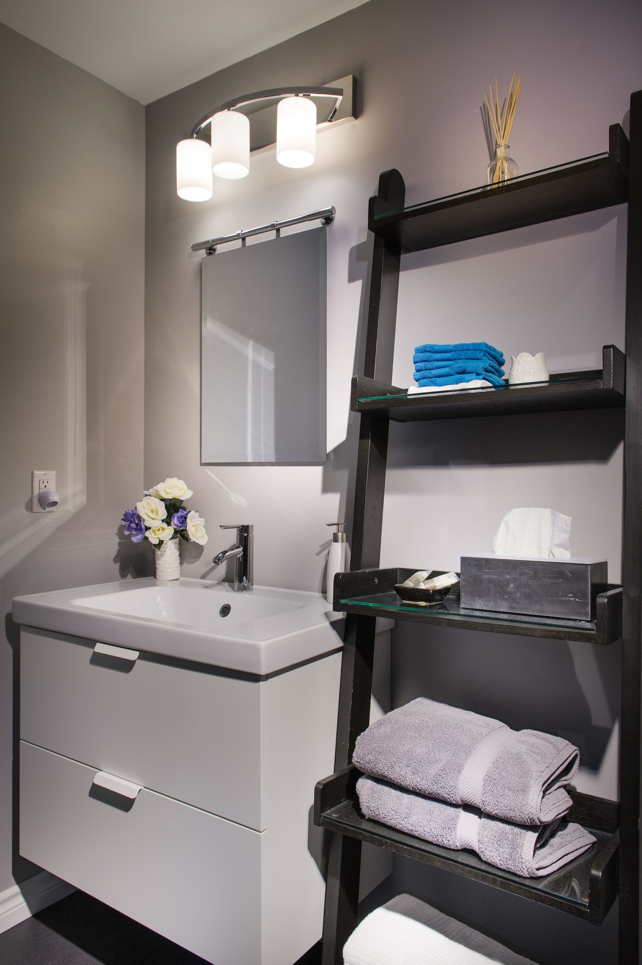 A modern bathroom with a white sink, a mirror, and a shelf with towels and toiletries.