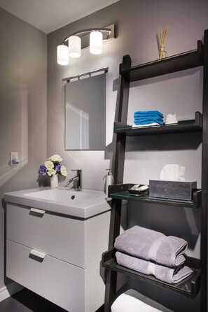 A modern bathroom with a white sink, a mirror, and a shelf with towels and toiletries. - Moncton Suites - 267 Universite Ave (Moncton)