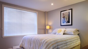 A bedroom with a large window, a bed with striped bedding, a bedside table, and a wall-mounted lamp. - Moncton Suites - 267 Universite Ave (Moncton)