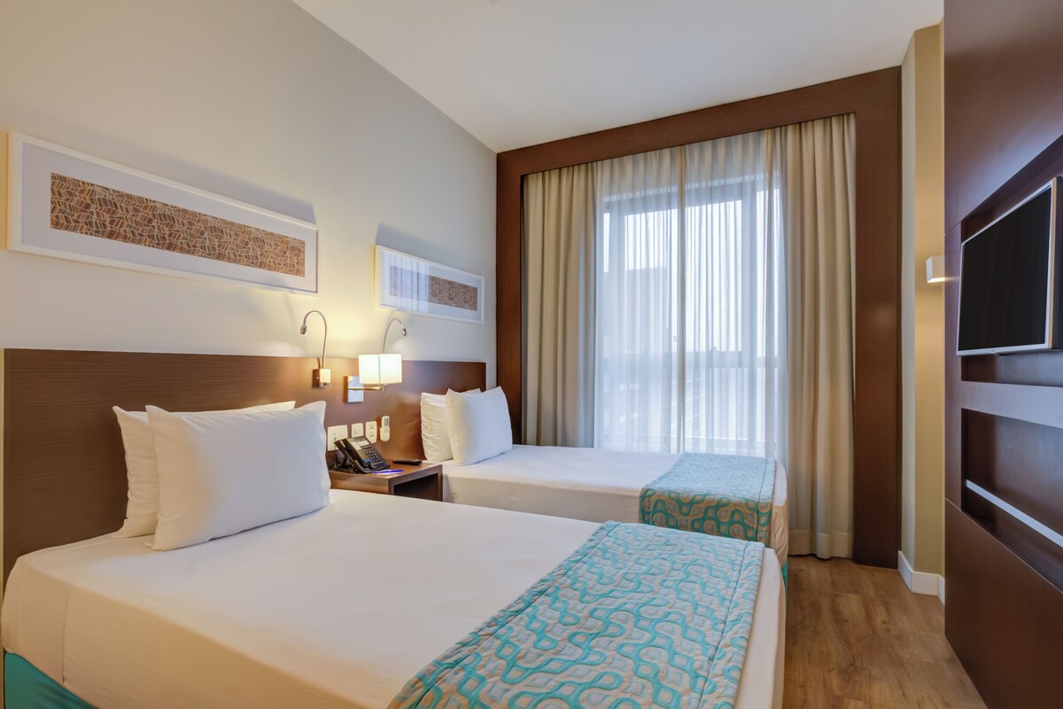 Een hotelkamer met twee bedden, een televisie, een bureau met telefoon en een raam met gordijnen.