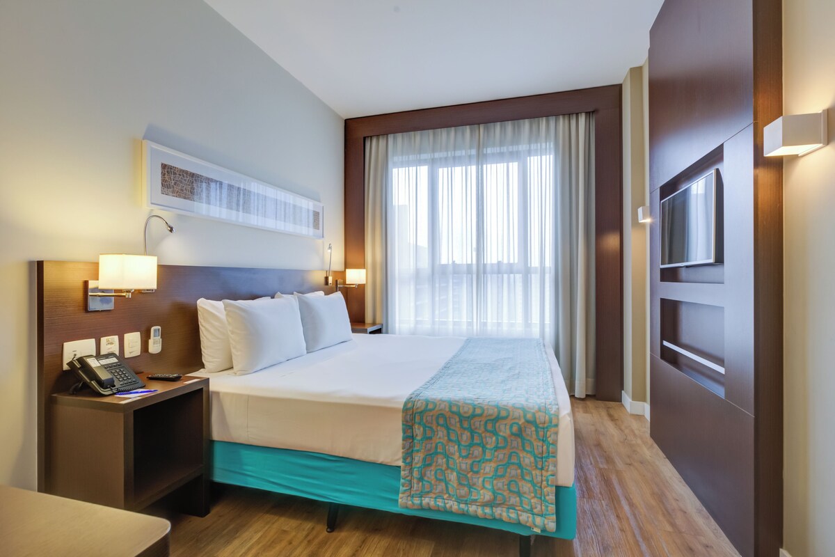 Een hotelkamer met een groot bed, een bureau met telefoon, een televisie en een raam met gordijnen.
