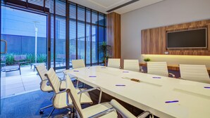 Een vergaderzaal met een lange tafel, witte stoelen en een groot raam met jaloezieën.