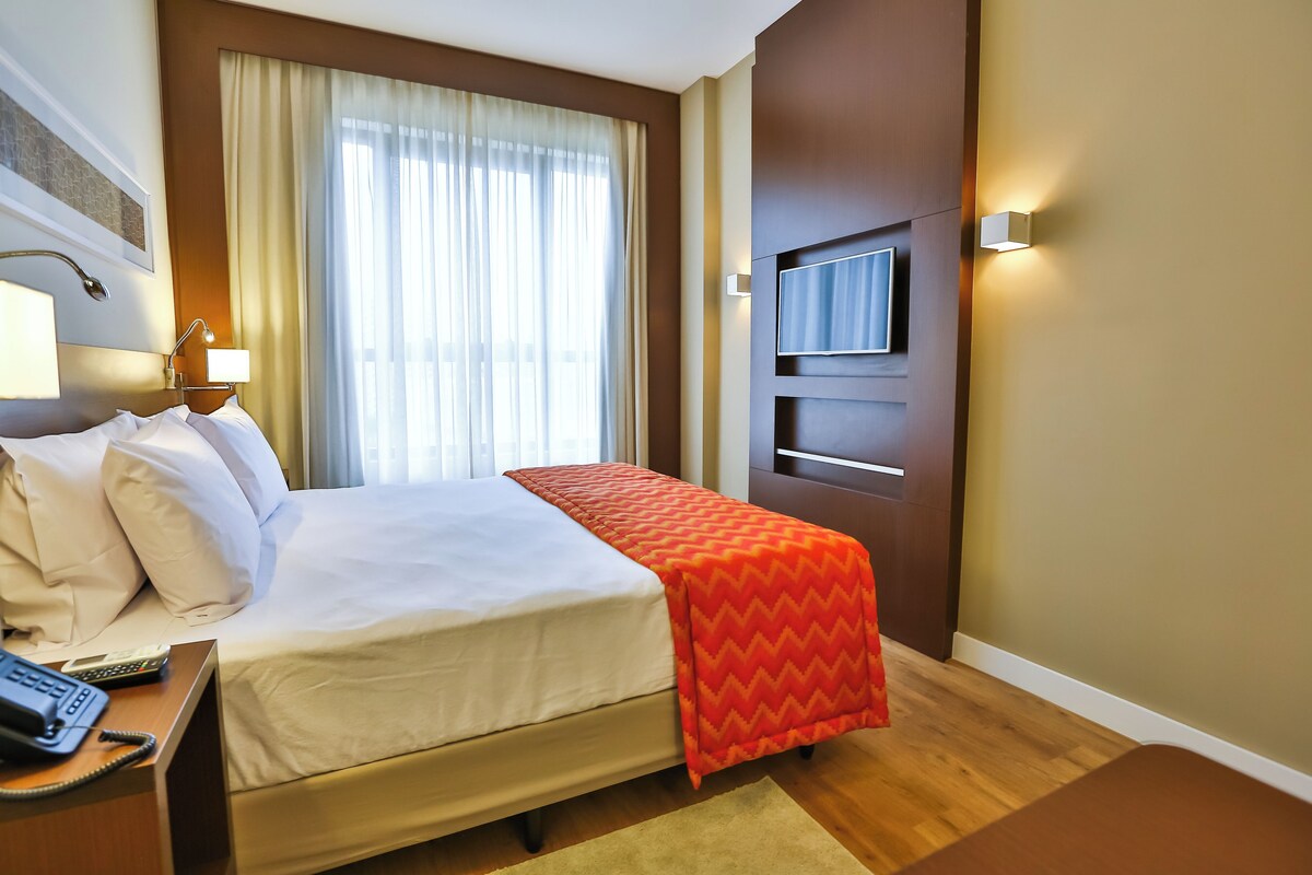 Hotelkamer met een bed, een aan de muur bevestigde televisie, een bureau met telefoon en een raam met gordijnen.