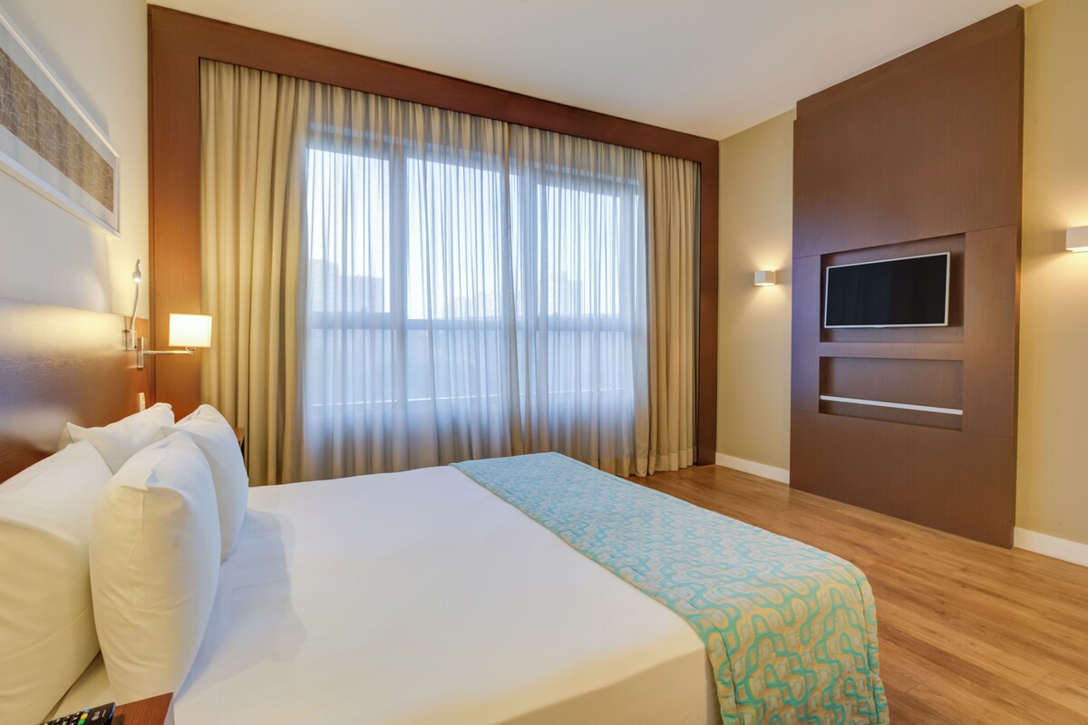 Een hotelkamer met een groot bed, een aan de muur bevestigde televisie en een raam met gordijnen.