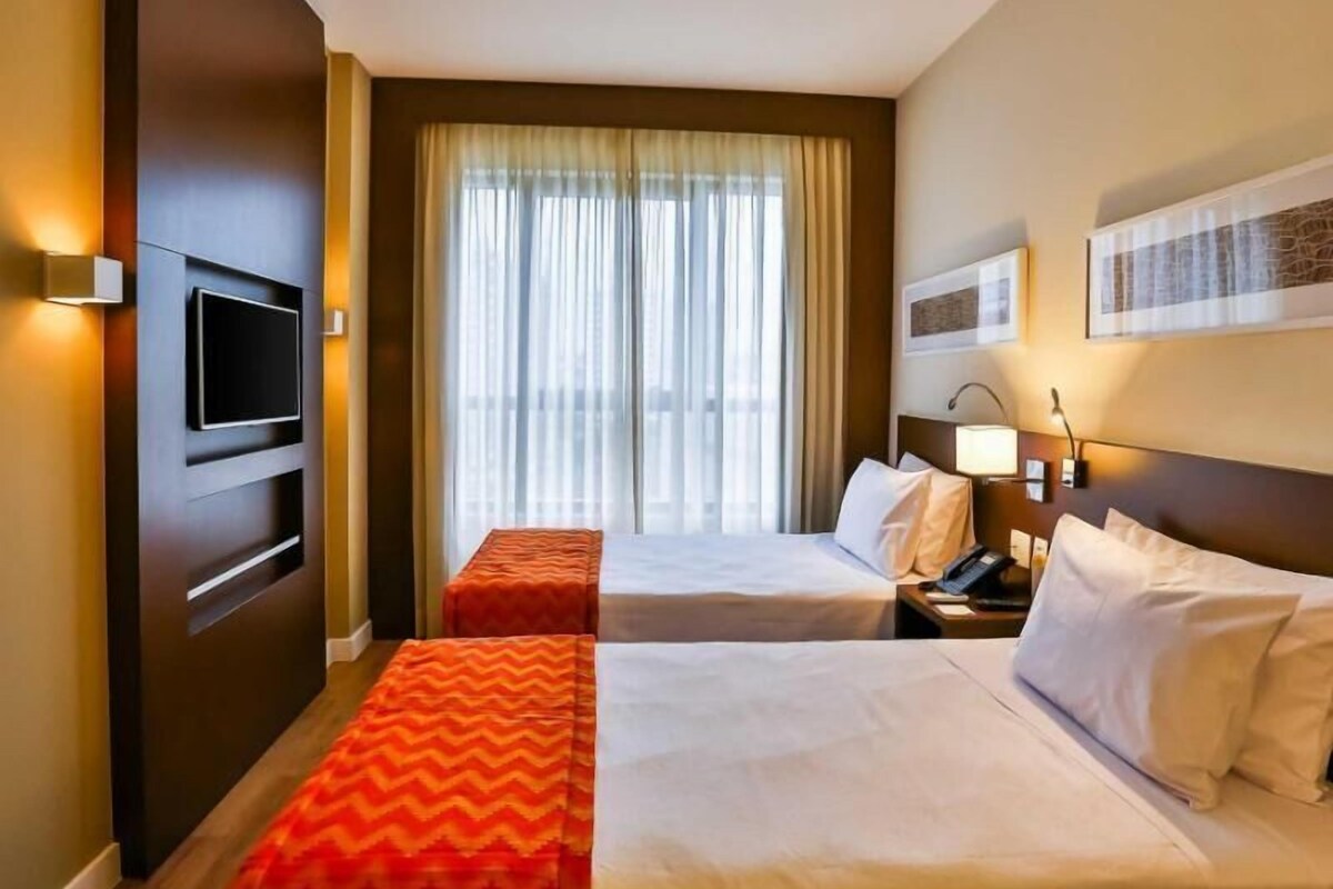 Een hotelkamer met twee bedden, een televisie, een bureau met lamp en een raam met gordijnen.