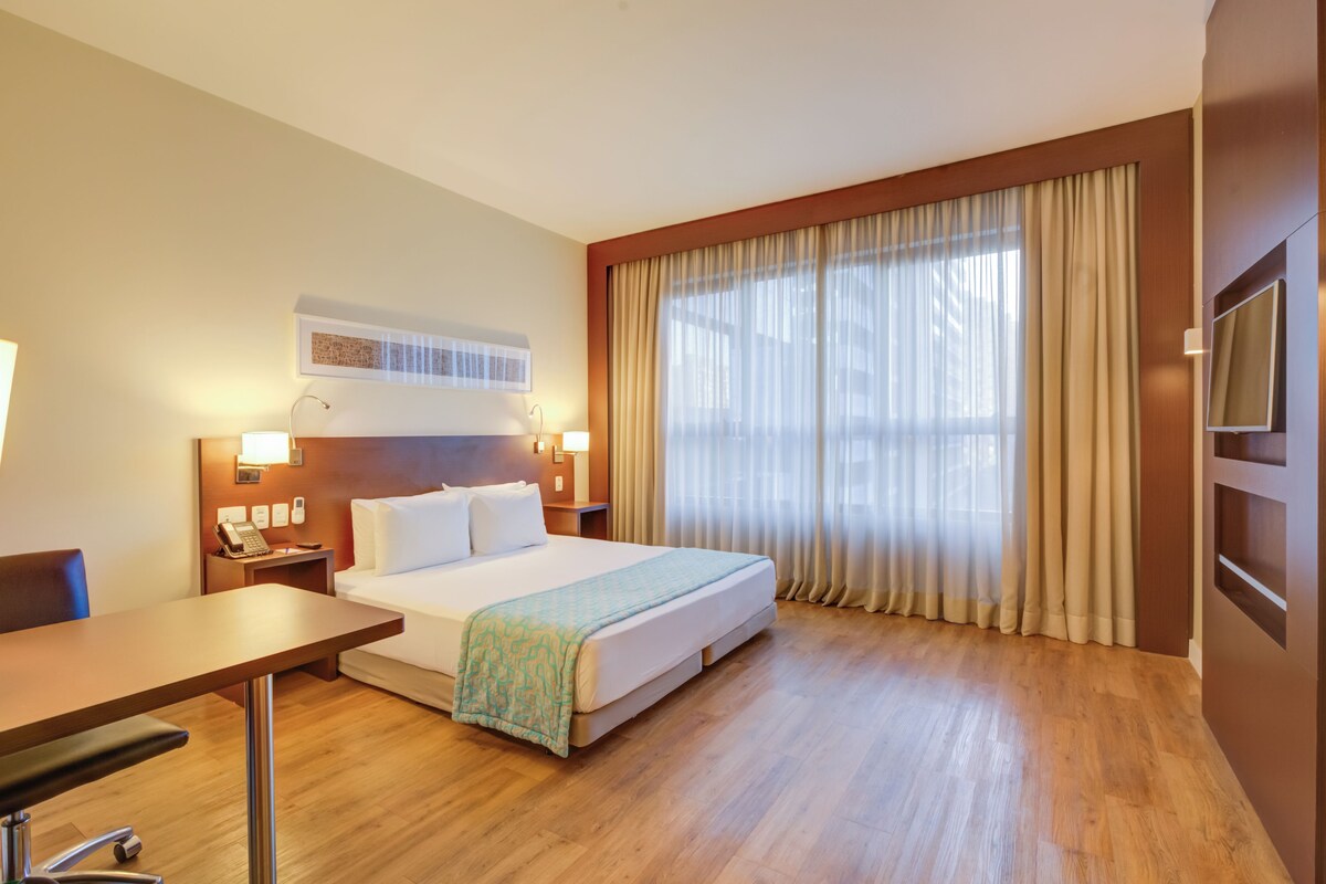 Een hotelkamer met een bed, nachtkastjes, een bureau en een groot raam met gordijnen.