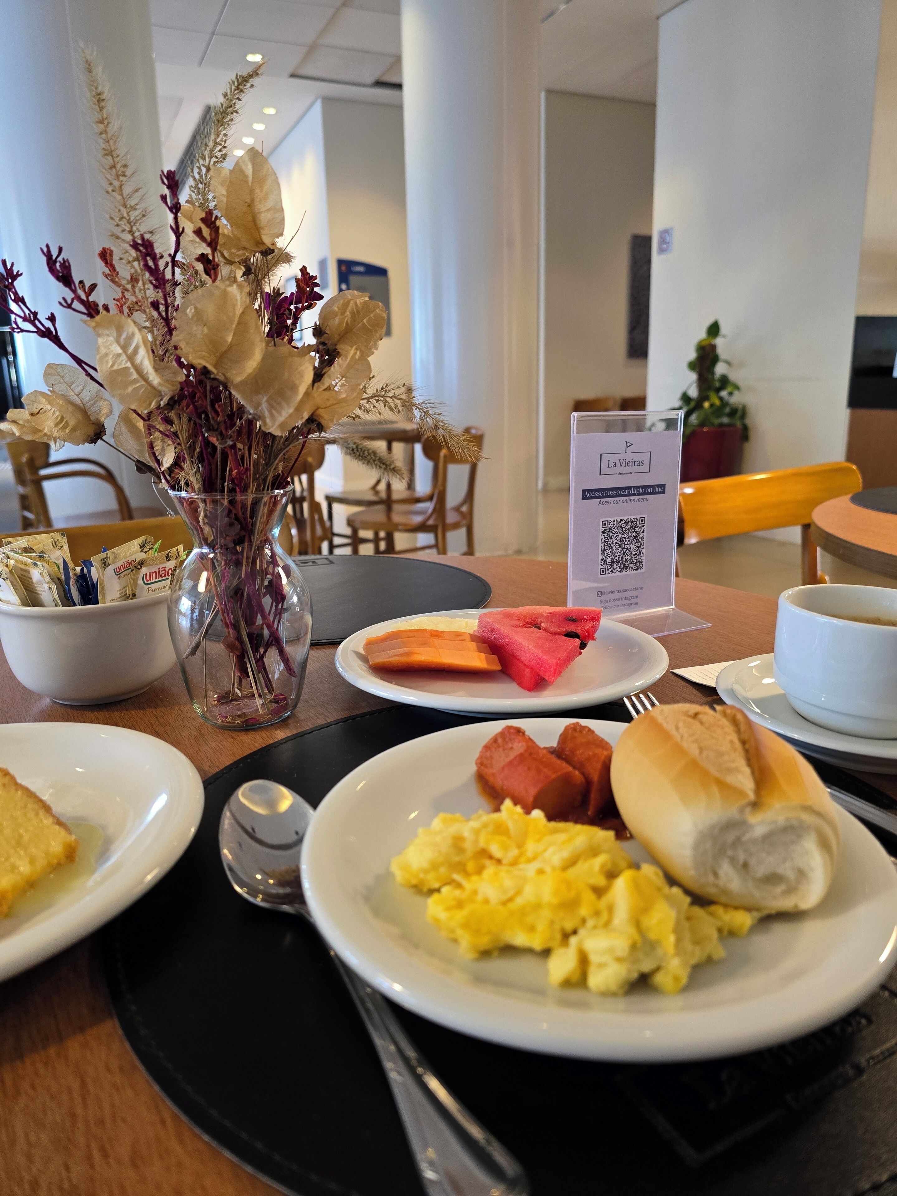 A breakfast setting with plates of food, a vase with dried flowers, a QR code sign, and a coffee cup.
