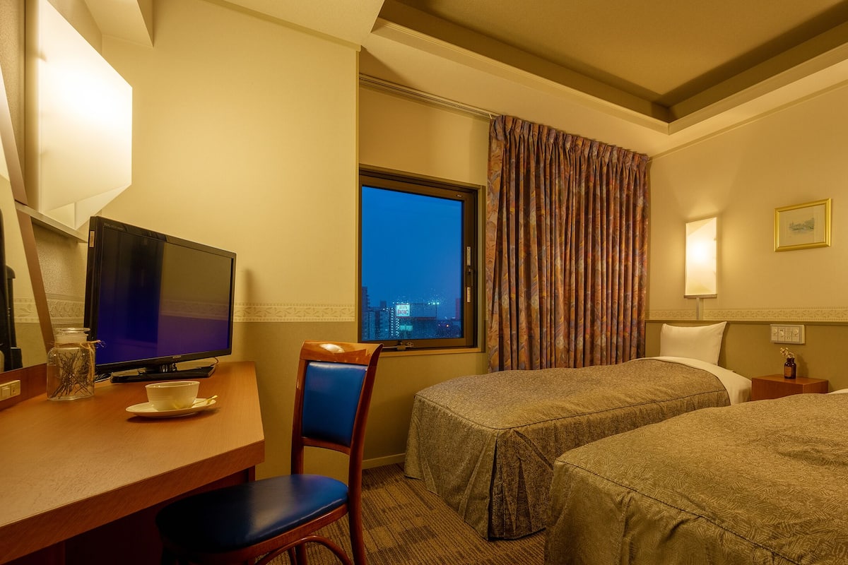 Habitación de hotel con dos camas, un escritorio con televisor, una silla y una ventana con cortinas.