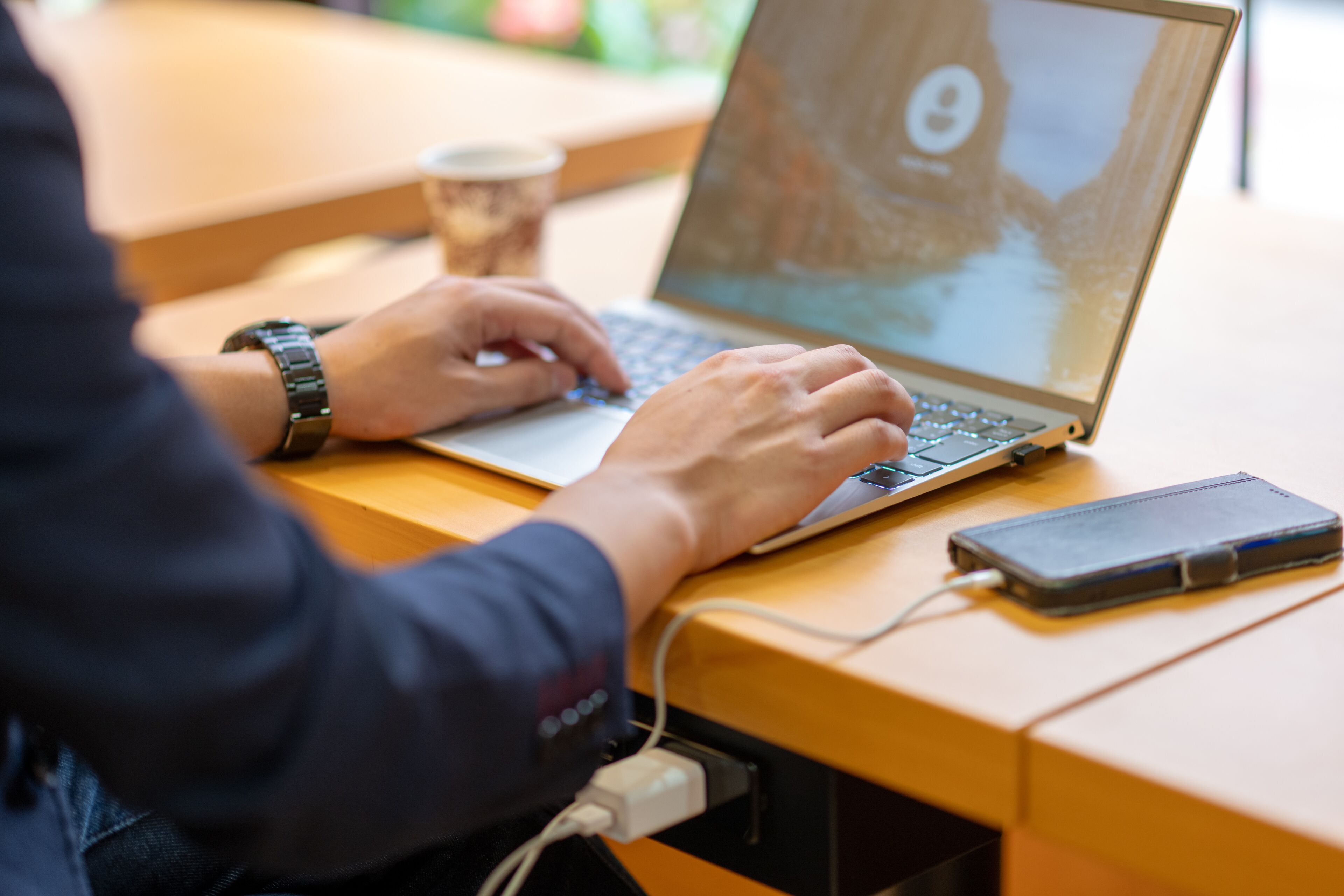 Una persona usando una computadora portátil sobre una mesa de madera, con un teléfono inteligente y una taza de café cerca.