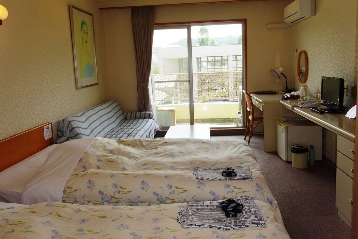 A hotel room with two beds, a desk, a chair, and a balcony with a view.