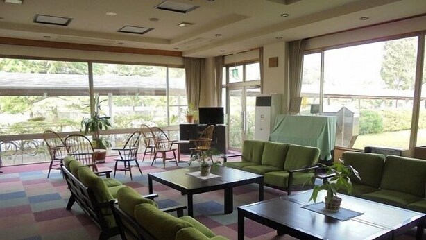 Un vestíbulo de hotel con sofás verdes, una mesa de centro y un televisor. Se observan grandes ventanales con vistas a la vegetación exterior.