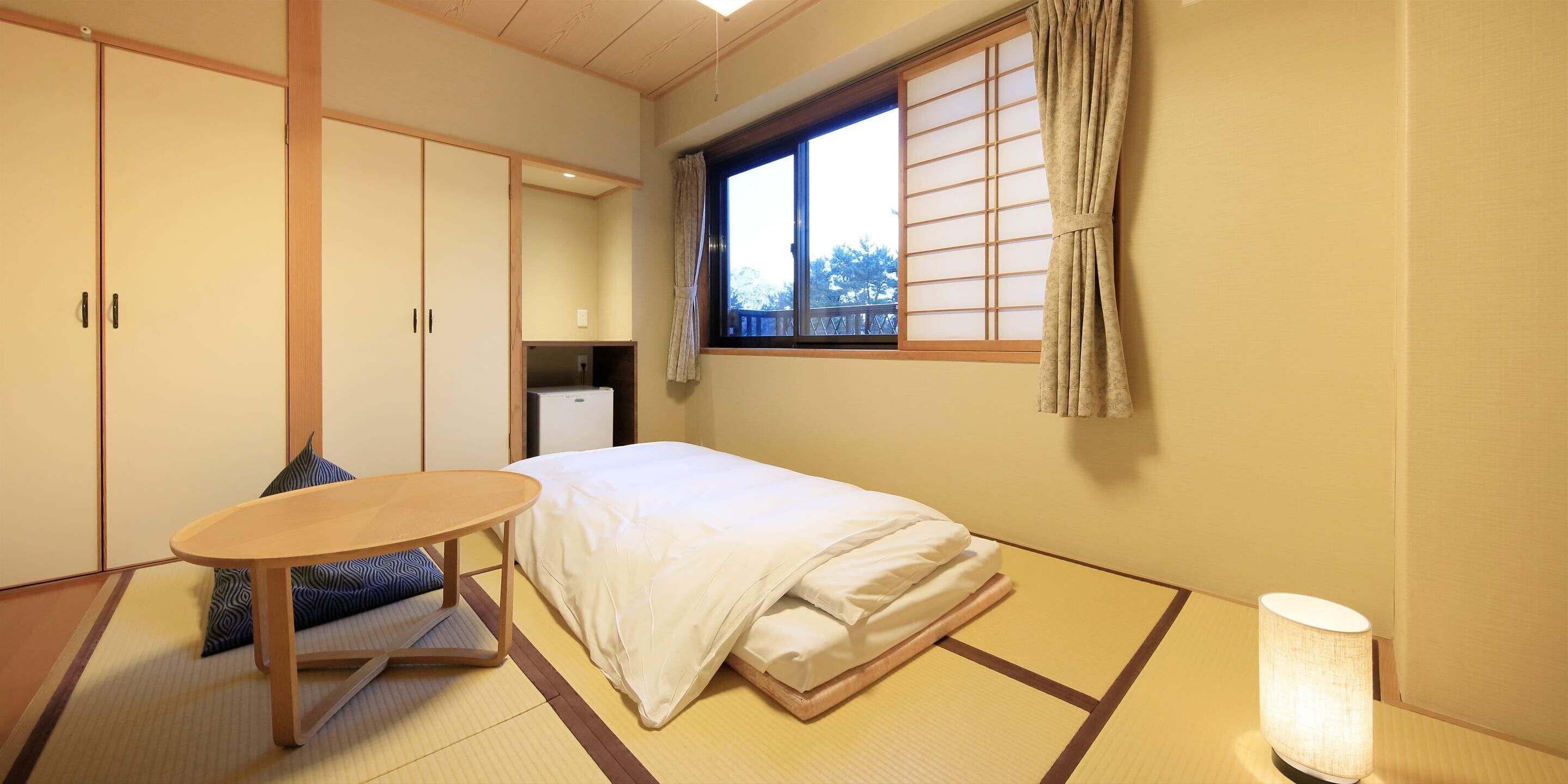 Perinteinen japanilaistyylinen huone, jossa on tatamimatto, matala sänky, pieni pöytä ja ikkuna, jossa on shoji-paperi-ikkunaluukut.