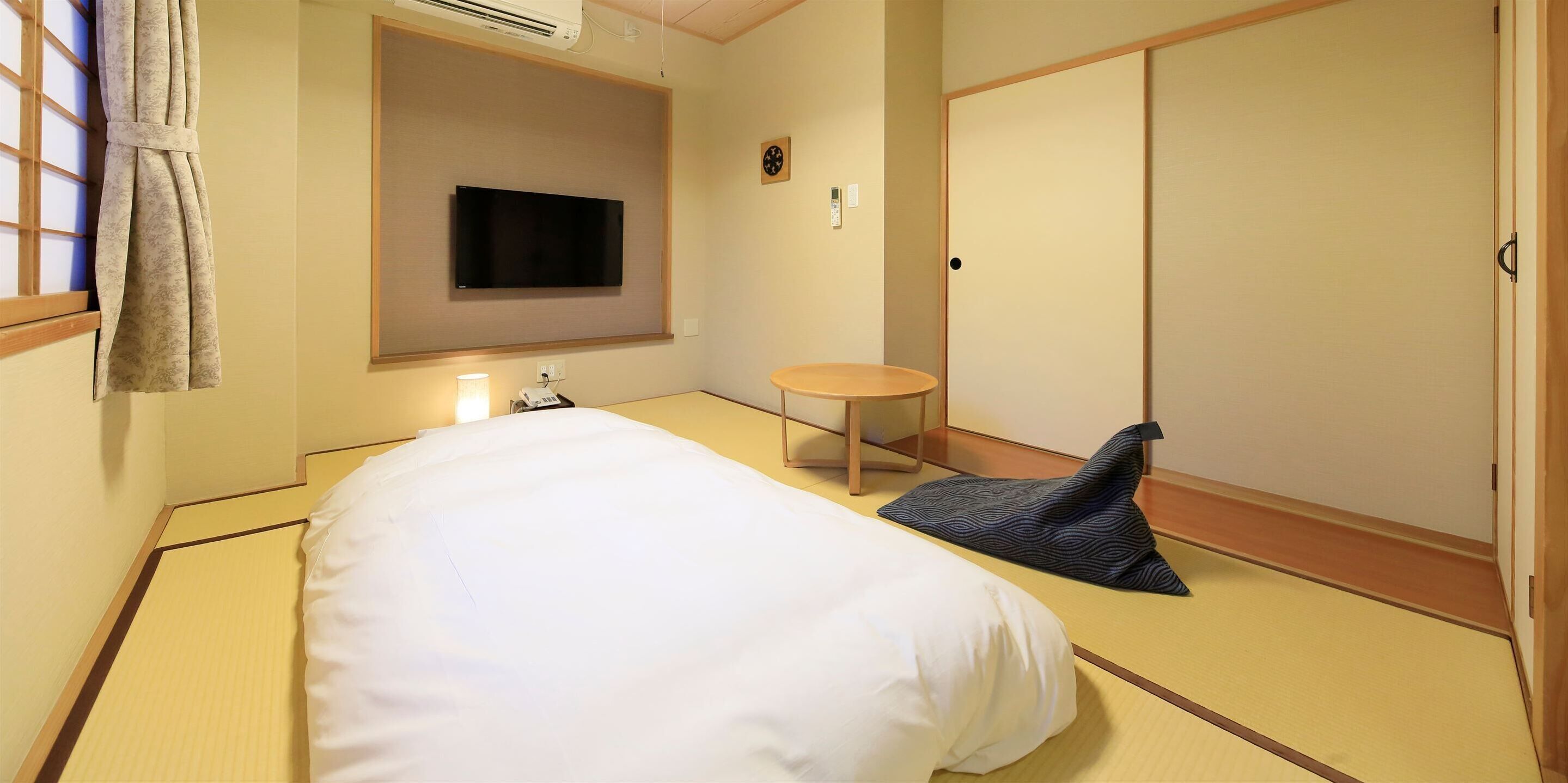Una habitación de estilo japonés tradicional con suelo de tatami, una cama con ropa de cama blanca, un televisor montado en la pared, una pequeña mesa redonda y una puerta corredera.
