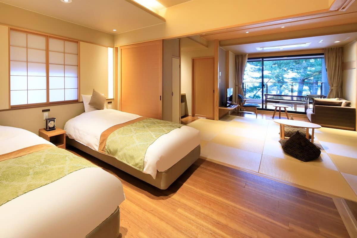 A hotel room with two beds, a sitting area with a table and chairs, and a large window offering a view of trees.