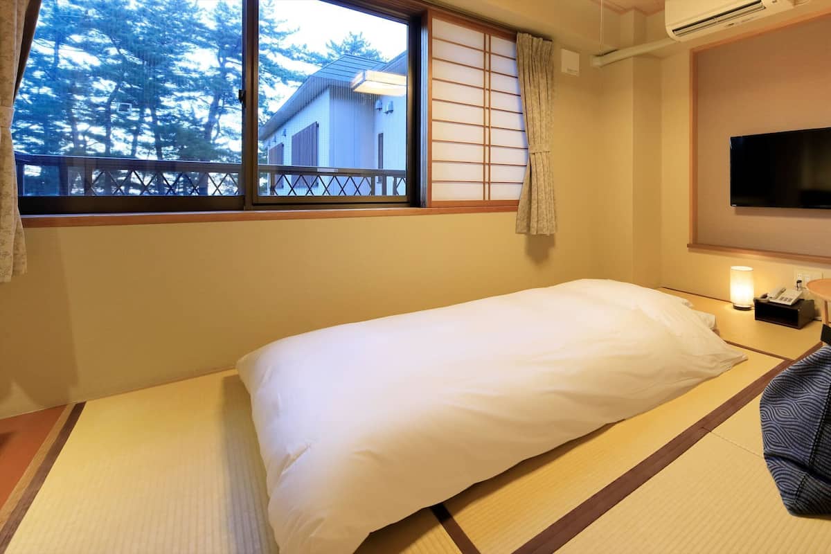 A hotel room with tatami flooring, a bed with white bedding, a television on the wall, and a small table with a lamp.