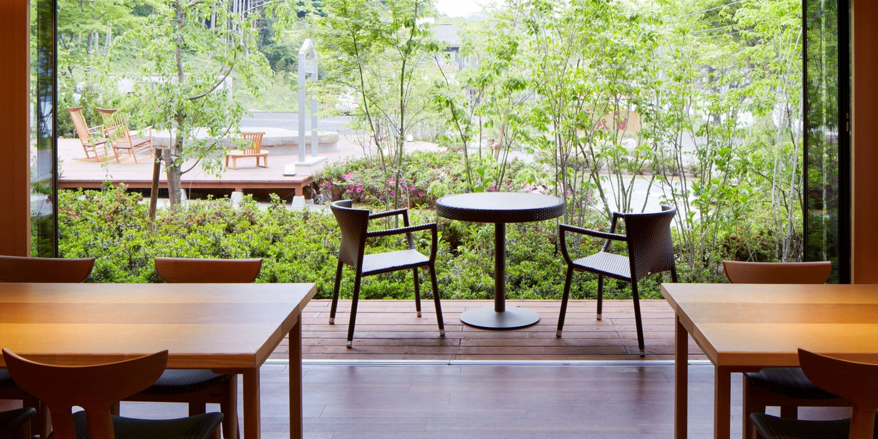 Un patio con mesas y sillas de madera, rodeado de abundante vegetación y vistas a la calle.