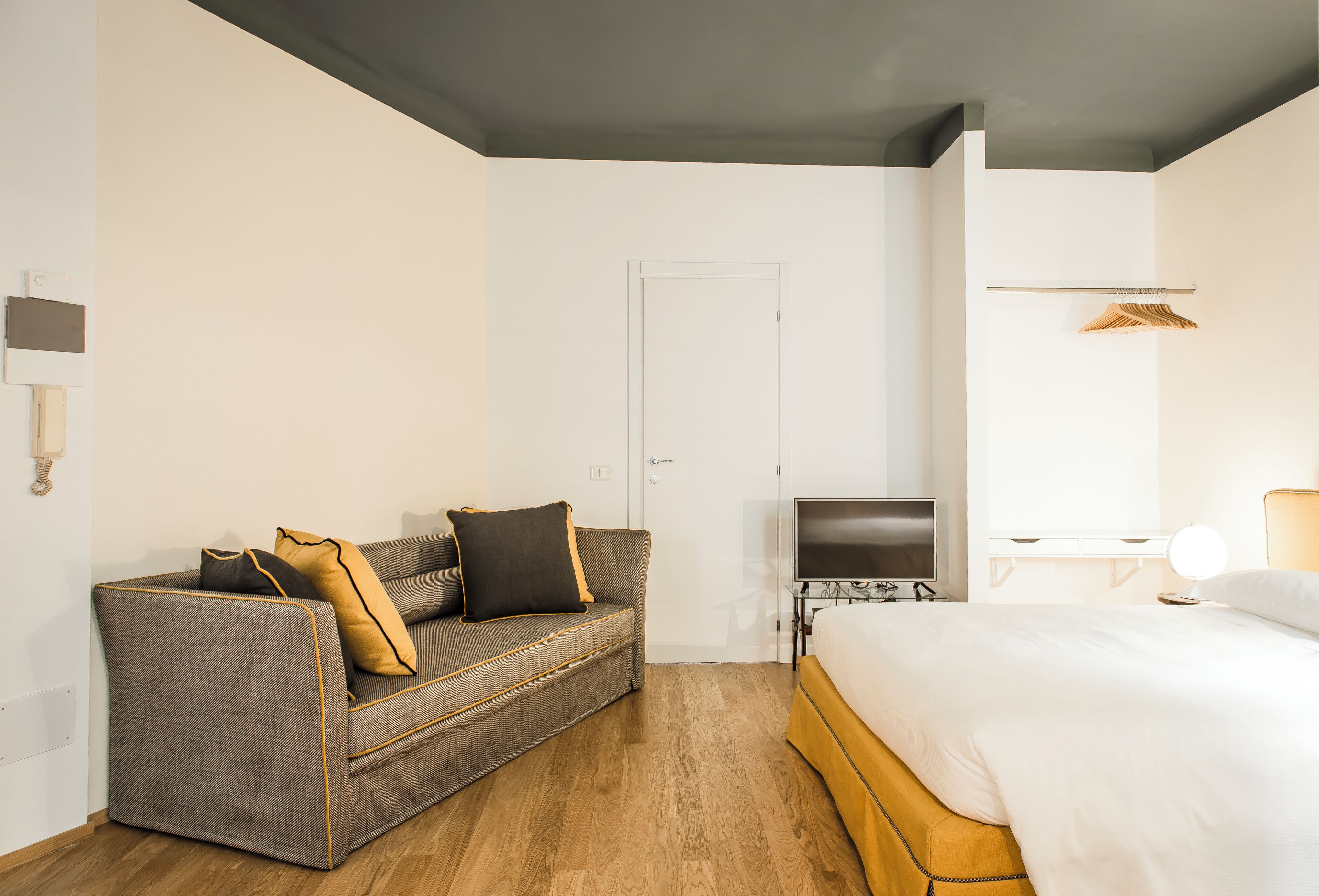 Habitació d'hotel amb un sofà gris, un llit, una televisió i una taula de nit amb una làmpada.