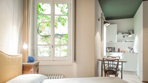 A bedroom with a large window, a bedside table with a lamp, a chair, and a view of a kitchen area. - Brera Apartments in Porta Romana (Milan)
