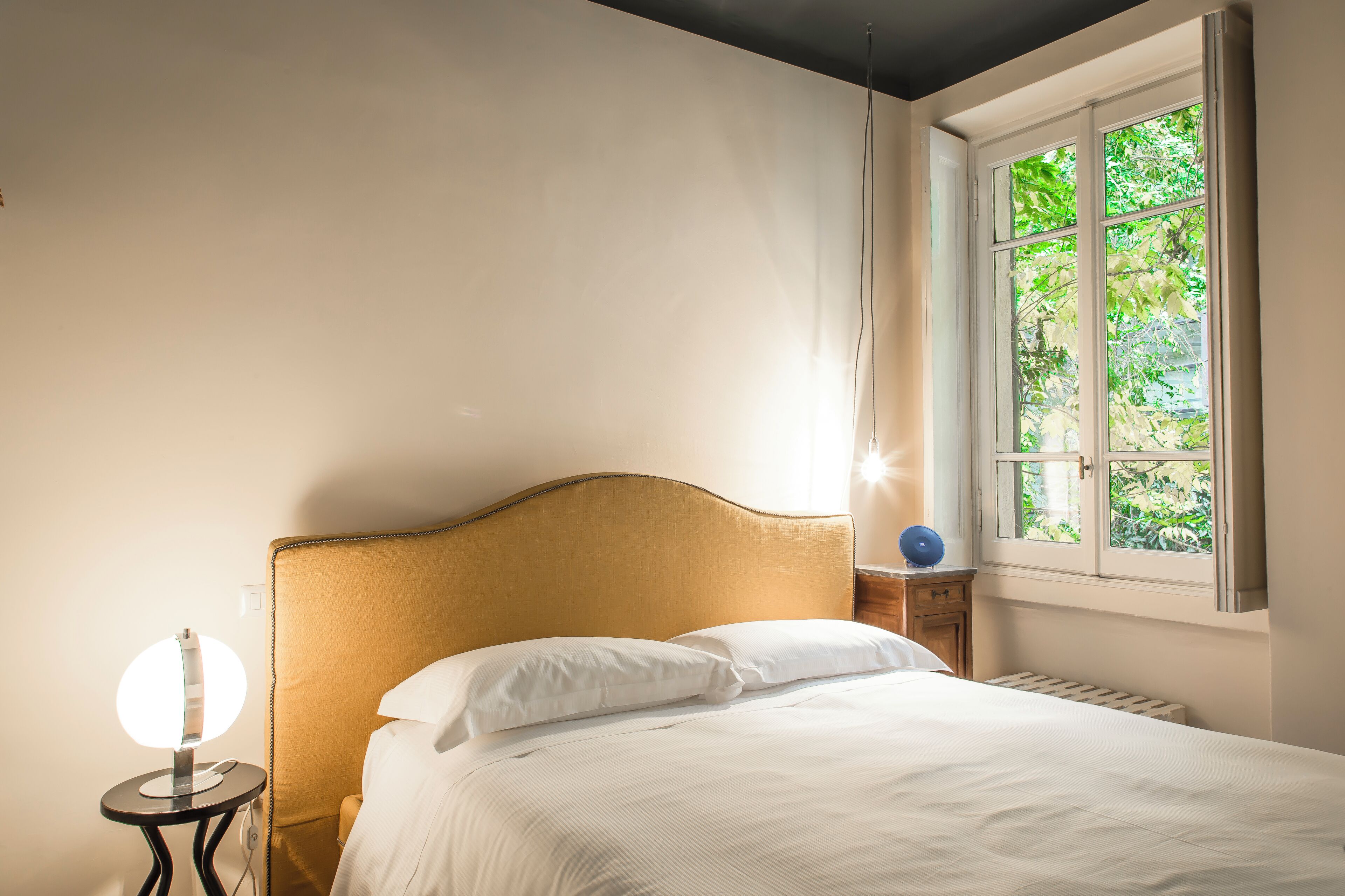 Una cama bien hecha con sábanas blancas, una mesita de noche redonda con una lámpara, y una ventana con vistas a la vegetación.