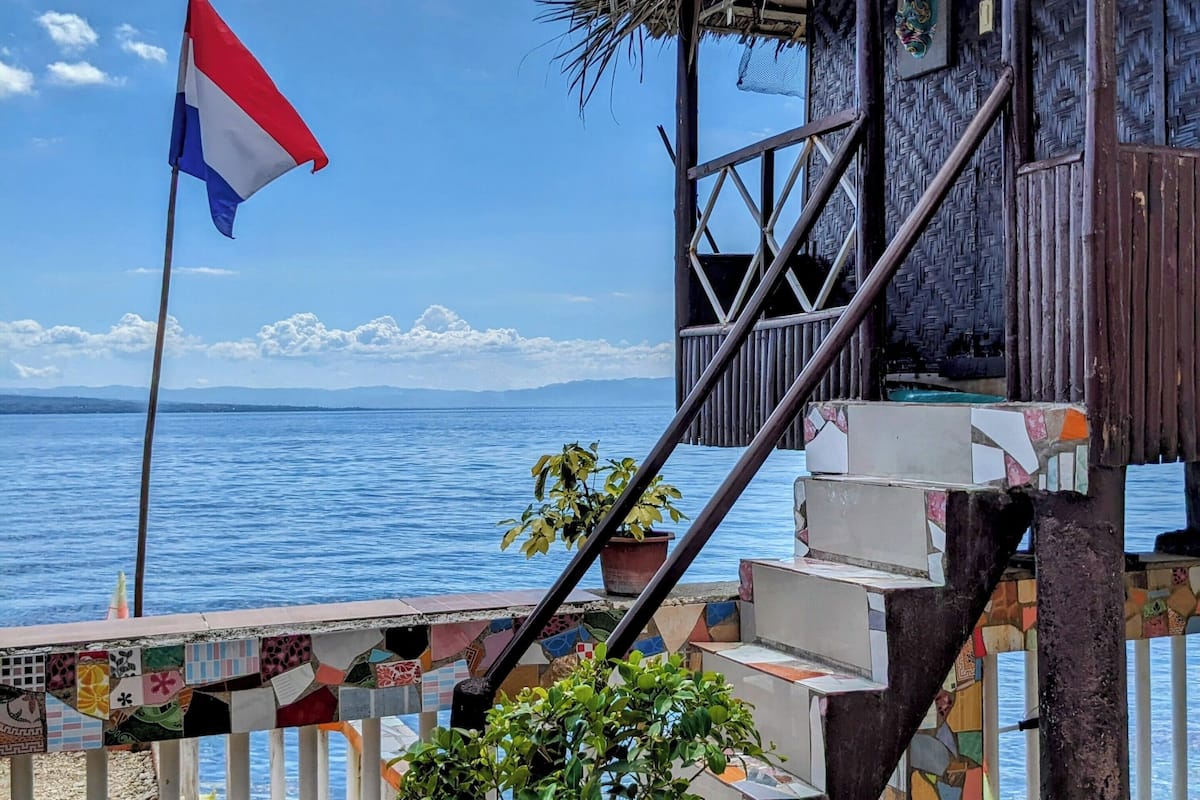 Un escalier en bois mène à une construction à toit de chaume, sur laquelle flotte un drapeau, et qui surplombe une étendue d’eau.