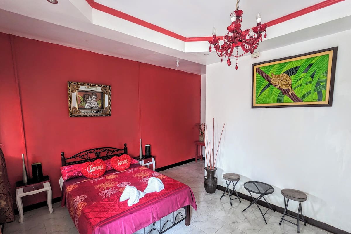 Une chambre avec un mur rouge, un lit avec une literie rouge, un lustre, un tableau encadré et une peinture verte.