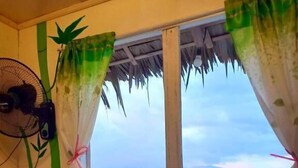 Une chambre avec un lit placé face à une fenêtre donnant sur un plan d’eau. Il y a deux ventilateurs au mur, une plante décorative et un oreiller sur le lit.