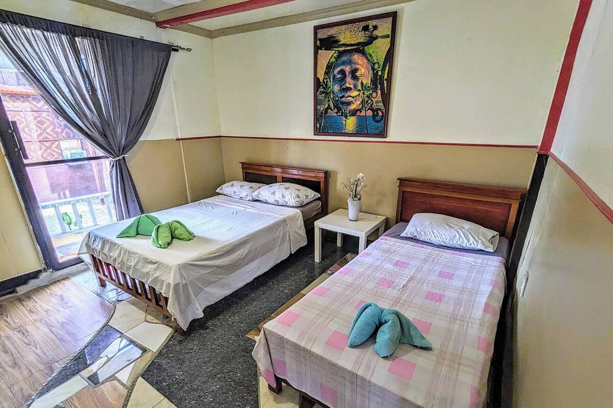 Une chambre avec deux lits, une petite table, une fenêtre avec des rideaux et un tableau au mur.
