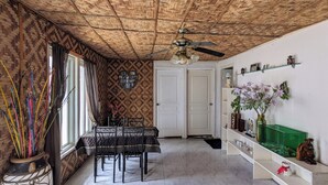 Une pièce avec un plafond en osier, une table à manger avec des chaises, un ventilateur de plafond et une étagère verte ornée d’objets décoratifs.
