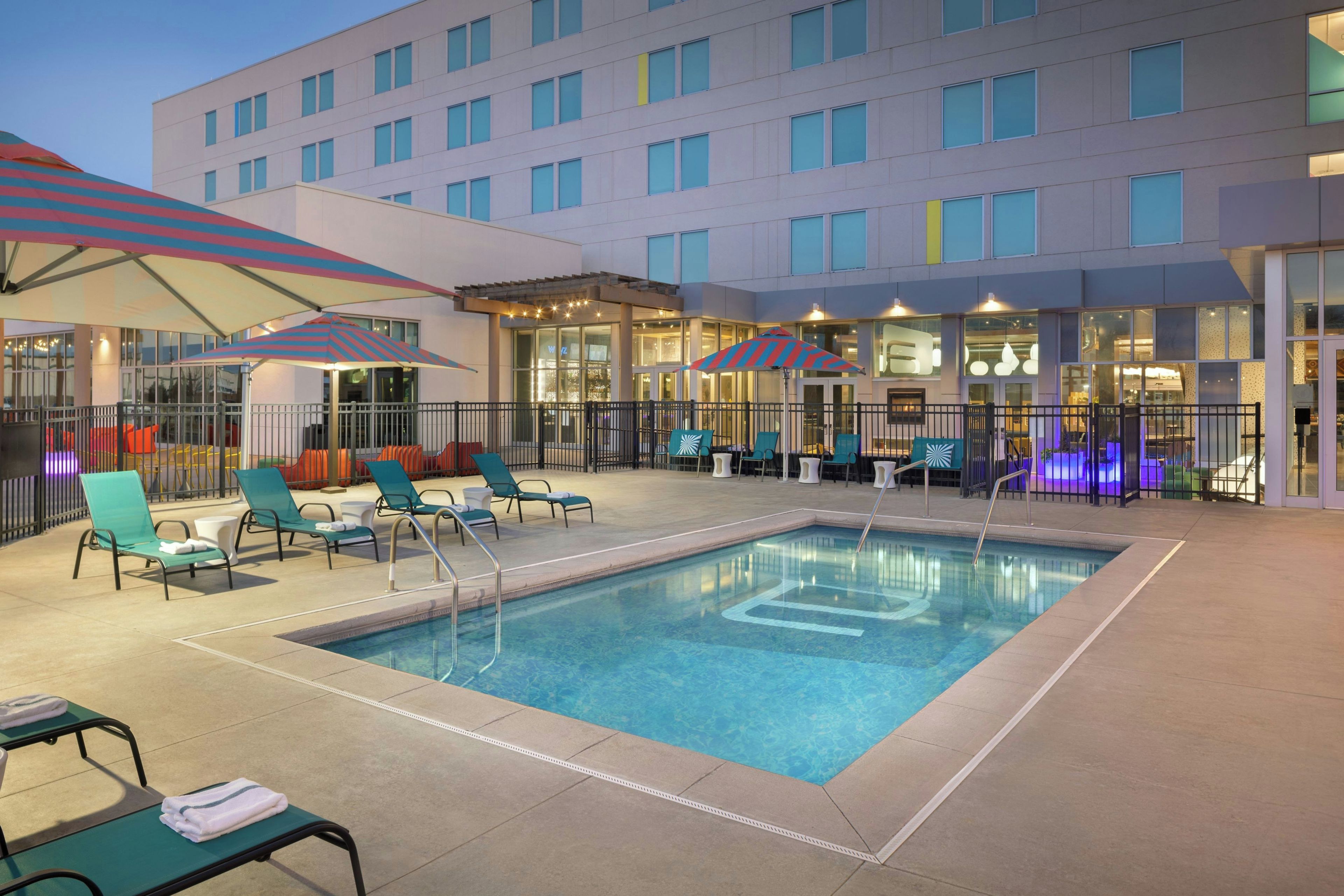 A hotel pool area with multiple lounge chairs and umbrellas, surrounded by a building with large windows.