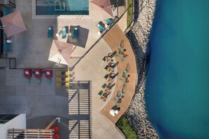 Vista aérea de un complejo turístico con piscina, tumbonas, sombrillas y una terraza de madera con vistas a una costa rocosa.