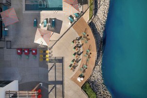 Vista aérea de un complejo turístico con piscina, tumbonas, sombrillas y una terraza de madera con vistas a una costa rocosa.