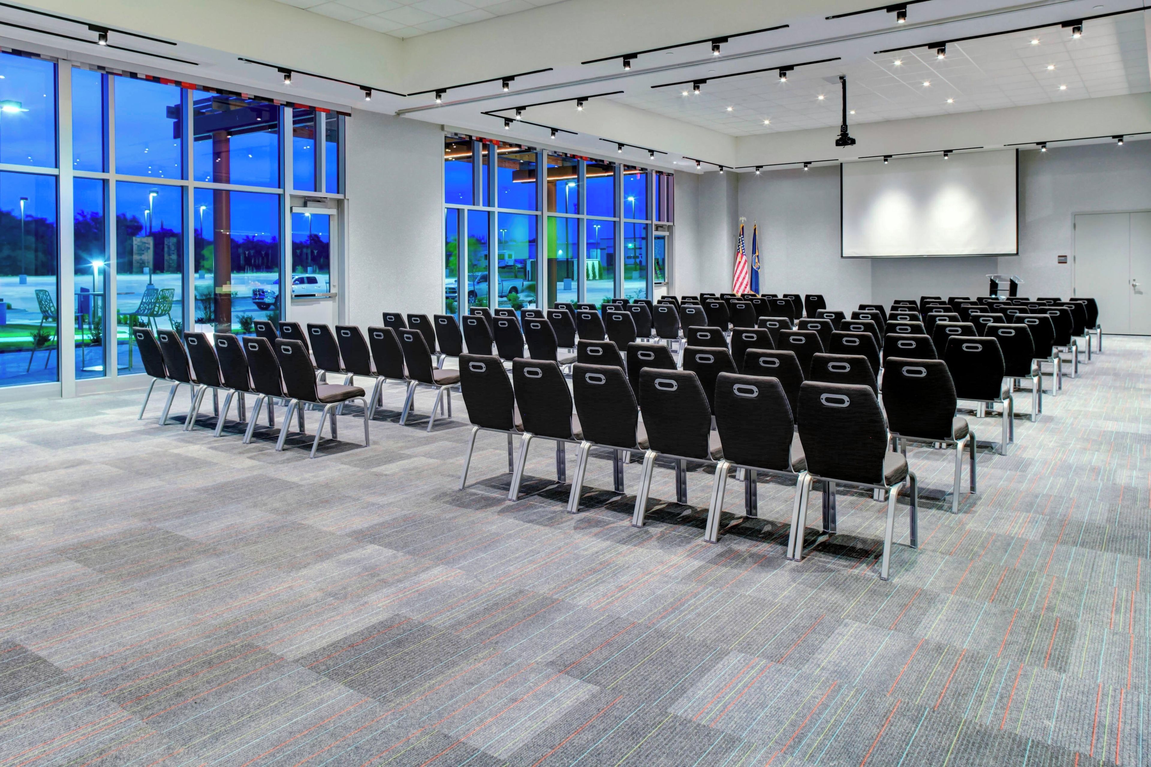 Un salón de conferencias con filas de sillas, ventanas amplias y una pantalla de proyección.